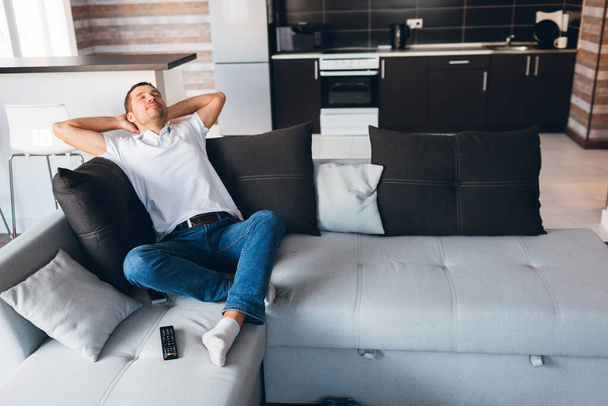 Young man watch tv in his own apartment. Guy enjoy his time in room. Sleeping joyful with hands under head. Lonely but happy. Rest and relax in new apartment. Daylight. - Photo, Image