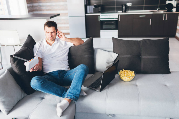 Mladý muž se dívá na televizi ve svém bytě. Chlápek sedí na gauči a telefonuje. Držte wi-fi router v ruce a podívejte se na něj. Přizpůsobení internetového zařízení. Otevřený notebook na gauči kromě. - Fotografie, Obrázek