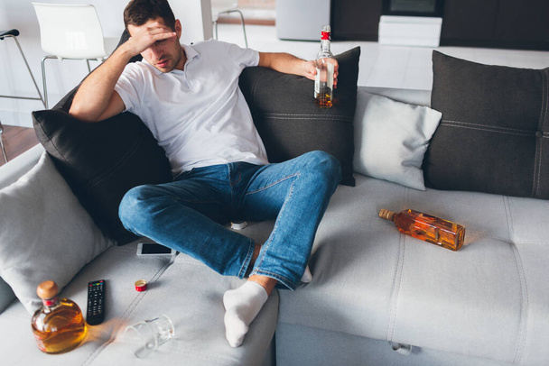 Young man watch tv in his own apartment. Alcoholic sit on sofa amoing opened bottles with whiskey. Suffer from hangover or headache. Alone with problems. Desparate and hopeless. - Photo, Image
