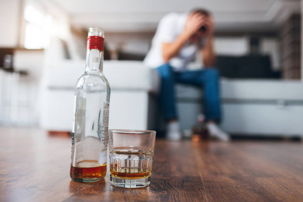 Young man watch tv in his own apartment. Picture with blurred background where guy sit on couch and suffer from hangover or headache. Alcoholic has troubles with drinking strong drinks. - Photo, Image