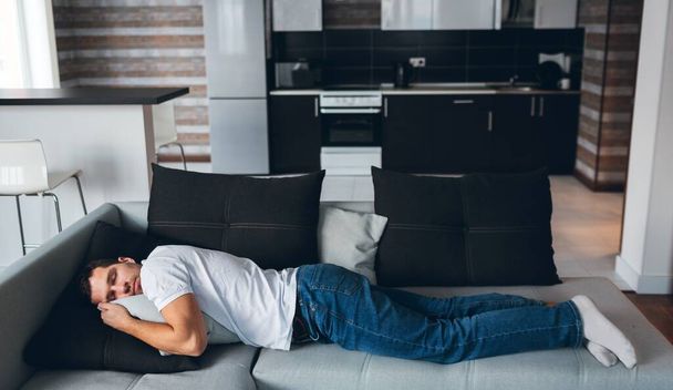 Young man watch tv in his own apartment. Guy lying in horizontal position on couch. Daydreaming with sweet dreams. Rest and relax. Calm peaceful man tired. - 写真・画像