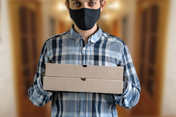 courier in a black mask and gloves with two cardboard boxes, close up of cardboard boxes against the background of a blurry man in a black mask - Φωτογραφία, εικόνα