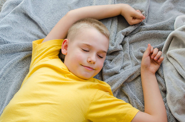 Mutlu sarışın çocuk evde uyanıyor. Sarı tişörtlü çocuk yumuşak gri battaniyenin üzerinde dinleniyor. Gündüz dinlenin. Evde kal. İzolasyon. Karantinaya alın. Üst görünüm - Fotoğraf, Görsel
