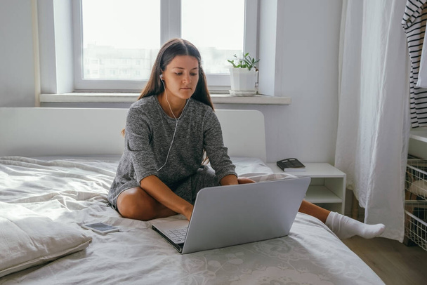 young woman in casual gray dress with earphones using laptop sitting on the bed in the bedroom at home - Foto, afbeelding