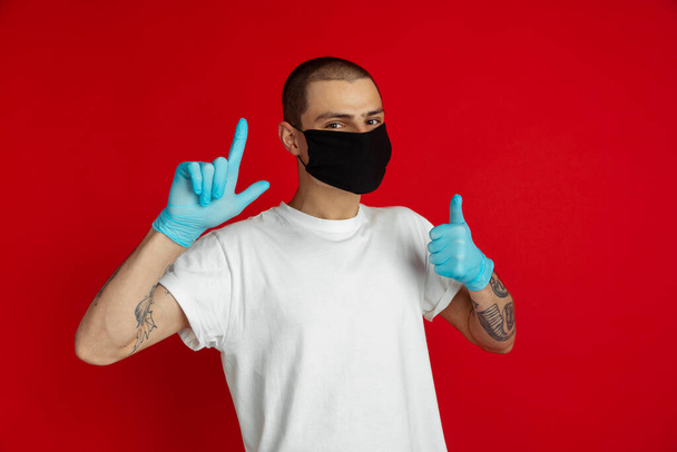 Caucasian young man in protective face mask and medical gloves on red studio background - pointing and showing thumb up - Photo, Image