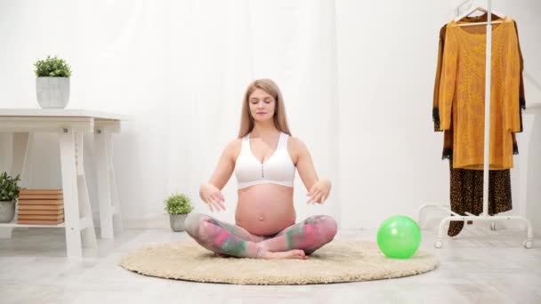 A young beautiful pregnant Caucasian woman sits on the soft carpet of the house. He is engaged in fitness and doing exercises. Sportswear. - Séquence, vidéo