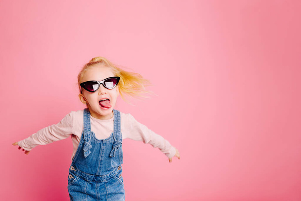Joven linda chica rubia olorosa está bailando y mostrando su lengua en gafas de sol retro sobre fondo rosa. Concepto de infancia feliz
. - Foto, Imagen