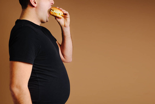 Fat man with big belly is eating hamburger. Abdominal pain from junk fat food. Unhealthy food. - Foto, afbeelding
