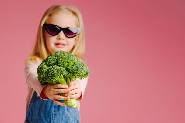 Young cute smiling girl in sunglasses is holding broccoli on pink background. Concept of happy childhood. Happy mother`s day or children`s day. - Foto, immagini