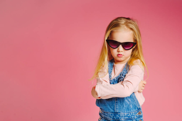 Young cute smiling girl in sunglasses is offended and angry on pink background. Concept of childhood. - Foto, imagen