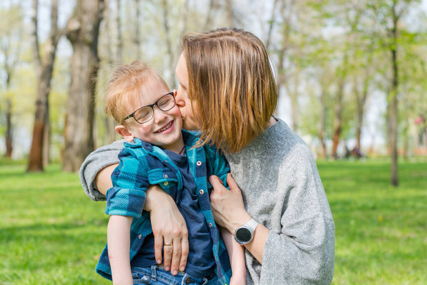 Mom kisses her little boy in glasses in the park on the grass in spring.  - Fotó, kép