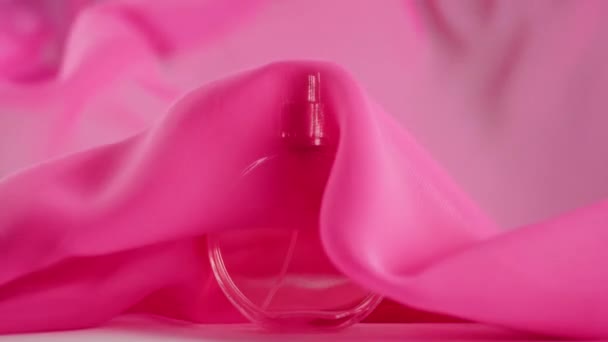 Ovale parfumflesje met roze parfum of etherische oliën staat op tafel. Roze stof fladdert rond en zwaait in de lucht rond de parfumfles. Concept van aroma en geur. Sluiten. - Video