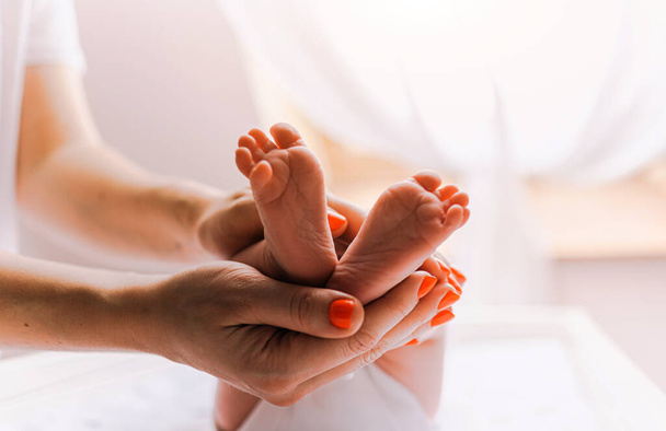 Close-up feet of a newborn baby. Young mother is holding baby feet in his hisands. Little infant is lying on the kids table. There is Newborn child on the baby changing table near window. - Foto, afbeelding
