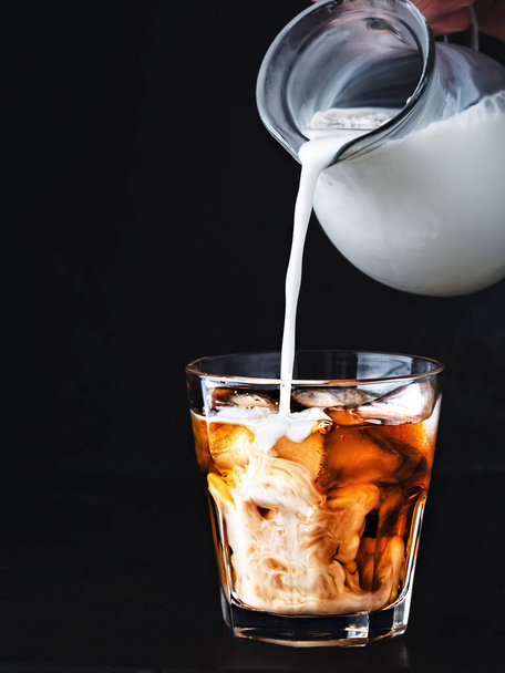 Iced coffee in a glass with ice and sugar syrup. Cream from a jug is poured into a glass. A refreshing and invigorating drink in summer in hot weather. Close-up, dark wood background. - Foto, afbeelding