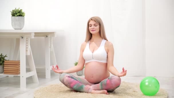 Молодая красивая беременная белая женщина сидит на мягком ковре дома. Занимается фитнесом и упражнениями. Спортивная одежда
. - Кадры, видео