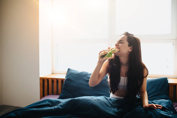 Νεαρή όμορφη γυναίκα στο πρωινό κρεβάτι στο σπίτι. Φαγητό και δάγκωμα σάντουιτς με πράσινα λαχανικά και νόστιμο ψωμί. Πρωινό στο σπίτι. - Φωτογραφία, εικόνα