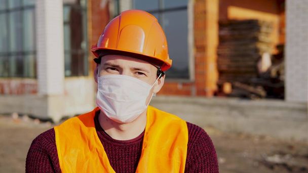 Portrait of male construction worker in medical mask and overalls on background of house under construction. - 写真・画像