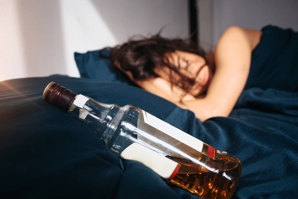 Young beautiful woman in morning bed at home. Sleeping after alcohol party. Bottle of whiskey on bed besides. Sun shines in room. - Photo, Image