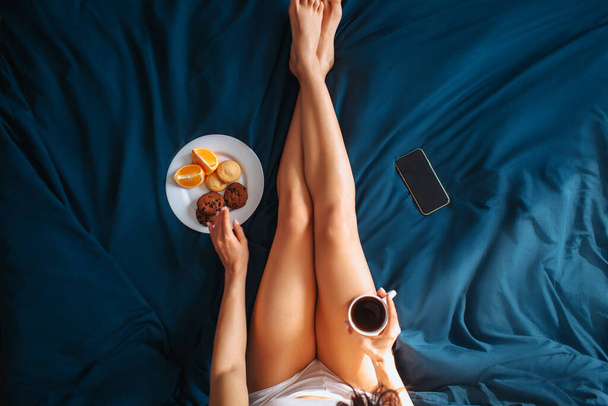 Jonge mooie vrouw in het ochtendbed thuis. Omhoog zicht op slanke goed gebouwde vrouwelijke benen en handen die een kop koffie vasthouden en de plaat bereiken met citrusvruchten, fruit en koekjes. Telefoon op bed. - Foto, afbeelding