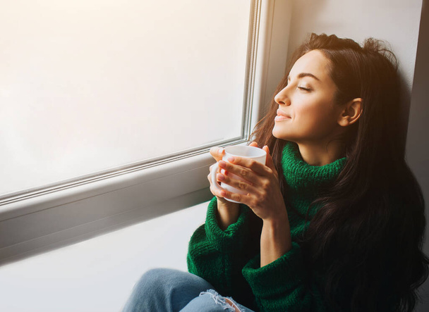 Idealny poranek Młoda brunetka siedzi na parapecie i trzyma filiżankę herbaty lub kawy w dłoniach. Kobieta modelka ubrana w zielony sweter siedzi w pobliżu i patrzy przez okno - Zdjęcie, obraz