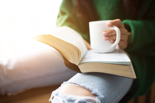 Τέλειο πρωινό Μια νεαρή μελαχρινή γυναίκα κάθεται σε ένα περβάζι και κρατά ένα βιβλίο και ένα φλιτζάνι τσάι ή καφέ στα χέρια της. Γυναικείο μοντέλο ντυμένο με πράσινο πουλόβερ μεγάλου μεγέθους. - Φωτογραφία, εικόνα