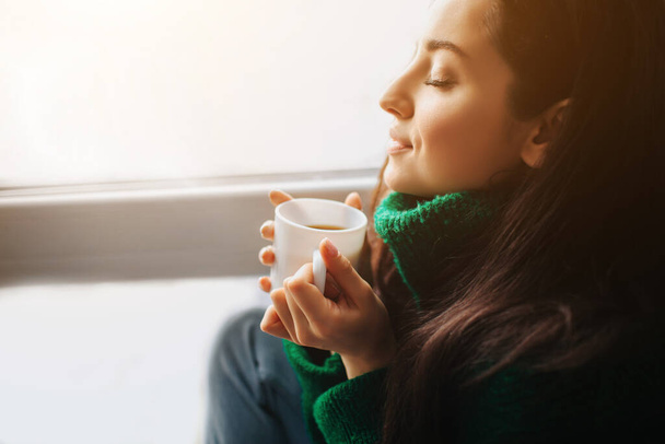 Tökéletes reggel Egy fiatal barna nő ül az ablakpárkányon, és egy csésze teát vagy kávét tart a kezében. Női modell öltözött zöld pulóver ül közel, és néz ki az ablakon - Fotó, kép