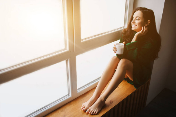 完璧な朝若いブルネットの女性は窓辺に座って、彼女の手に紅茶やコーヒーを持っています。女性モデル服を着て緑のセーターで座っていると窓の外を見て - 写真・画像