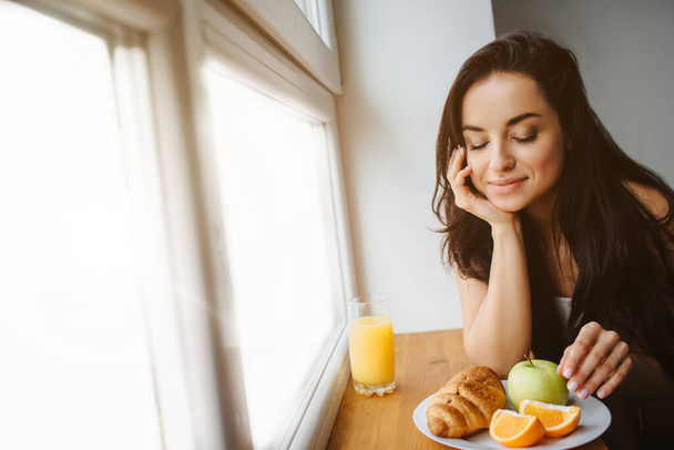白い下着の良い朝の若いブルネットの女性は木製の窓辺で朝食を持っています。若い女性モデルが食べる。新鮮なオレンジやジュース、クロワッサン、果物、ホワイトプレート上のリンゴがあります - 写真・画像