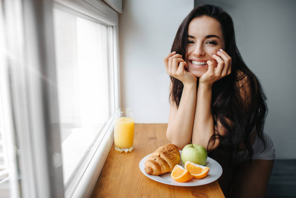 Καλημέρα νεαρή μελαχρινή γυναίκα με λευκά εσώρουχα τρώει πρωινό σε ξύλινο περβάζι. Ένα νεαρό θηλυκό μοντέλο τρώει. Υπάρχει φρέσκο πορτοκάλι ή χυμό, κρουασάν, φρούτα, μήλο σε λευκό πιάτο - Φωτογραφία, εικόνα