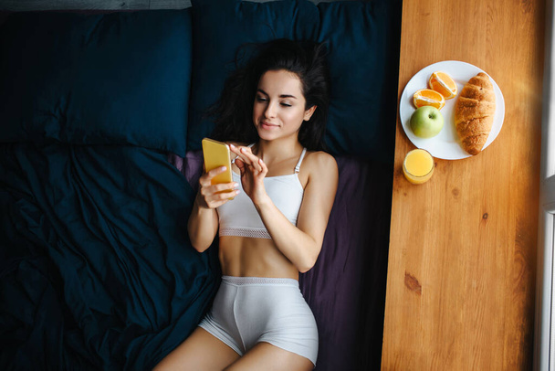 Young brunette woman in white underwear has breakfast on a wooden windowsill. She is using a phone. top view. There is fresh orange or juice, croissant, fruit, apple on a white plate - Fotografie, Obrázek