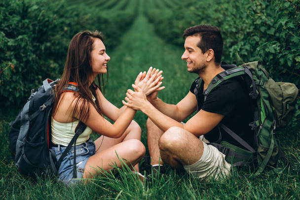 Homme et femme avec des sacs à dos sur le dos assis étreignant sur l'herbe verte entre les groseilles. Promenade sur les plantations de groseilles à la campagne
. - Photo, image