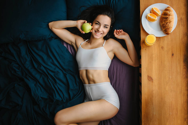 Good morning Young brunette woman in white underwear has breakfast on a wooden windowsill. A young female model eats. There is fresh orange or juice, croissant, fruit, apple on a white plate - Φωτογραφία, εικόνα