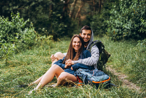 Woman with long hair and man sitting on the grass in the forest with backpacks. Have fun in nature, hugging and kissing. - Fotografie, Obrázek