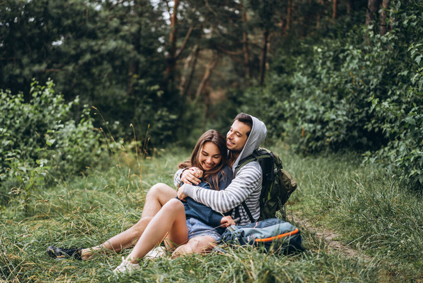 Woman with long hair and man sitting on the grass in the forest with backpacks. Have fun in nature, hugging and kissing. - Фото, зображення