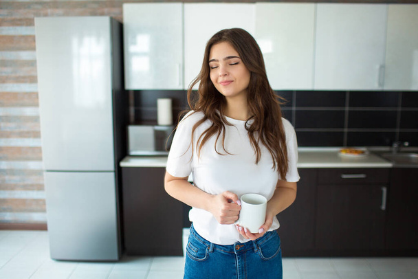 Egy fiatal nő a konyhában a karantén alatt. Pozitív vidám álmodozó modell csésze a kezében és csukott szemmel. Egyedül a konyhában. Napfény.. - Fotó, kép
