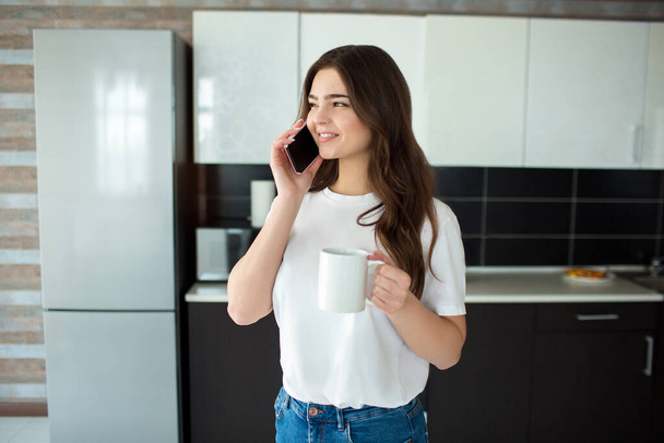 Молодая женщина на кухне во время карантина. Разговаривать по смартфону и улыбаться. Современные технологии в карантинный период. Работа в Интернете
. - Фото, изображение