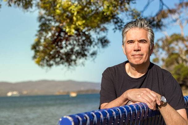 Portrait d'un homme mature de 57 ans avec une caméra aux cheveux gris assis sur un banc sur une plage à l'extérieur, confiant, détendu
 - Photo, image