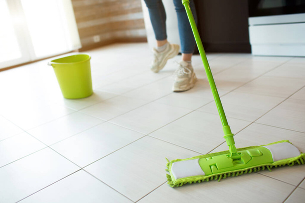 隔離中の台所の若い女性。緑のモップで床を掃除します。低カットビュー。床をよりきれいにする。後ろに水の入った緑のバケツ. - 写真・画像