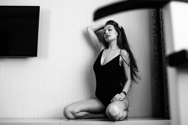 Young beautiful girl with good body shapes, half-naked, in a black bodysuit, sitting on a table in a hotel in front of the window - Fotó, kép