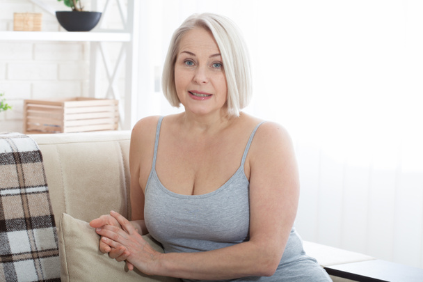 Active beautiful middle-aged woman smiling friendly and looking in camera in living room. Womans face close up. Realistic images without retouching with their own imperfections. Selective focus. - Foto, Bild