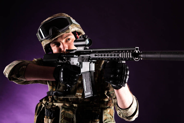 Soldier with gun is looking through the scope on violet background. Concept of war. Veterans, comrades, soldiers. Man in uniform. - Photo, Image