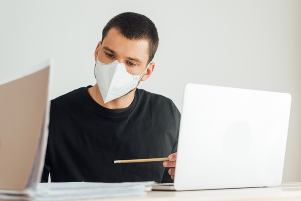 selective focus of freelancer in medical mask looking at folder near laptop  - Fotografie, Obrázek