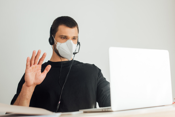 selective focus of operator in medical mask and headset waving hand while having video call at home - Foto, Bild