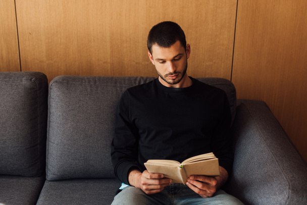 gutaussehender und bärtiger Mann liest Buch im Wohnzimmer  - Foto, Bild