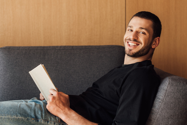 hombre sonriente y barbudo sosteniendo libro en la sala de estar
  - Foto, Imagen