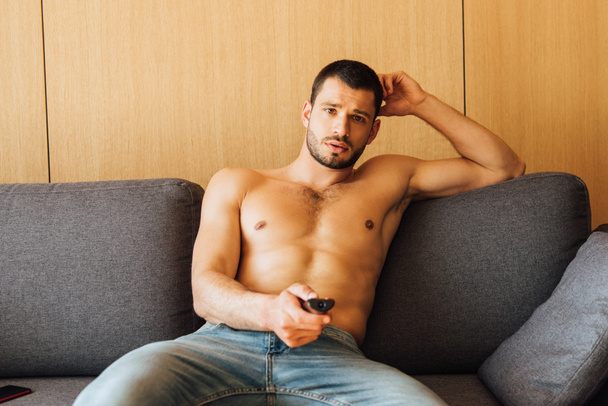Muž bez košile v džínách sledující film a držící dálkové ovládání  - Fotografie, Obrázek