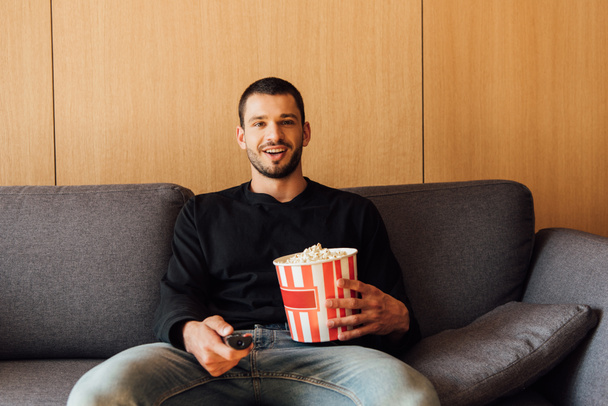 happy and bearded man holding remote controller and popcorn bucket  - Fotó, kép