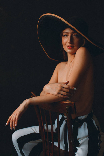 Young beautiful girl with good body shapes, nude, against a black wall, she has wide striped pants and a large black hat with brim - Zdjęcie, obraz