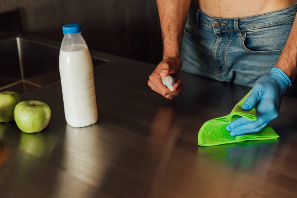 cropped view of man holding bottle with antibacterial liquid and rag near bottle of milk and apples on wooden table - Foto, imagen