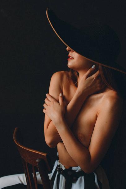 Young beautiful girl with good body shapes, nude, against a black wall, she has wide striped pants and a large black hat with brim - Photo, image